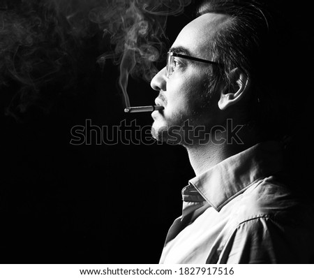 Portrait of brutal strong man business shark in official shirt and glasses stands with cigarette in his mouth, smokes over dark background. Side view. Fashion for men and stylish casual look concept