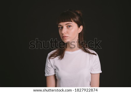 Studio portrait of a pretty brunette woman in a white blank t-shirt, half-turned, against a plain black background, looking at camera