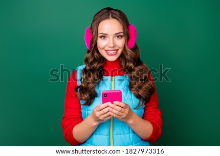 Photo of charming lovely young lady hold telephone chatting boyfriend share common pictures social network wear pink ear warmers blue vest red pullover isolated green color background