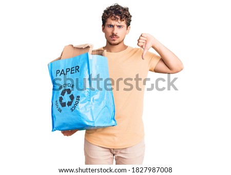 Young handsome man with curly hair holding recycling wastebasket with paper and cardboard with angry face, negative sign showing dislike with thumbs down, rejection concept 