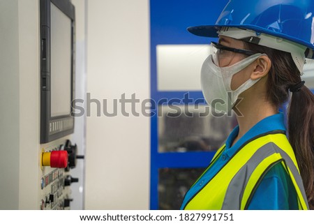 portrait of asian female factory tecnician with safety vest and helmet wearing face mask to protect coronavirus covid-19 working in factory control room at CNC machine control panel