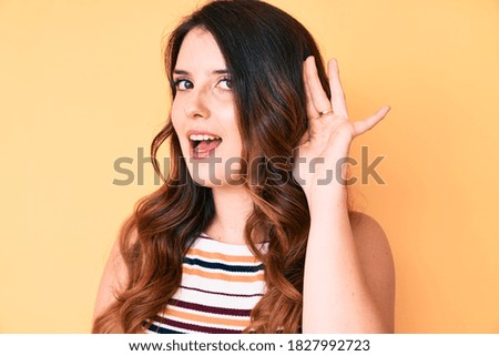 Young beautiful brunette woman over yellow background smiling with hand over ear listening and hearing to rumor or gossip. deafness concept. 