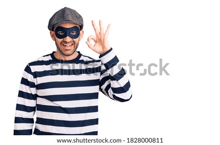 Young handsome man wearing burglar mask smiling positive doing ok sign with hand and fingers. successful expression. 