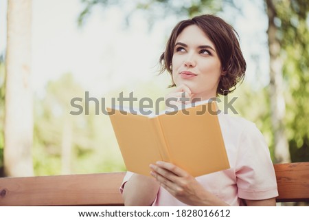 Photo of adorable pretty dreamy young lady look side hand touch chin hold book think imagine story plot get new information facts exam preparing wear pink t-shirt sit bench park outdoors Royalty-Free Stock Photo #1828016612