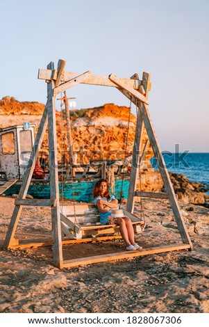 Romantic evening at sunset by the sea, a young woman in a dress with a bouquet of flowers is sitting on a wooden swing holding her hat