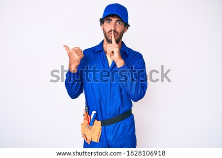 Handsome young man with curly hair and bear weaing handyman uniform asking to be quiet with finger on lips pointing with hand to the side. silence and secret concept. 
