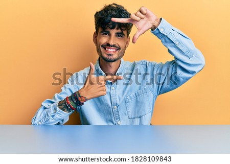 Young hispanic man wearing casual clothes sitting on the table smiling making frame with hands and fingers with happy face. creativity and photography concept. 