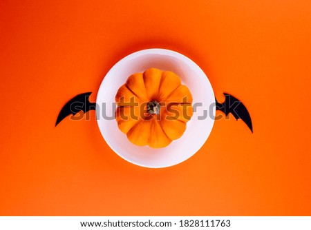 Whole raw orange pumpkin on white round plate with hand carved black bat wings in center of horizontal orange paper background. Copy space. Creative flat lay. Funny image with Halloween symbols.
