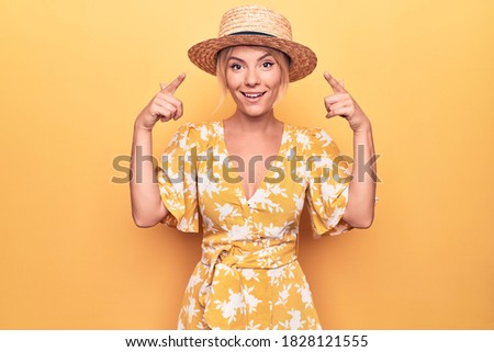 Beautiful blonde woman on vacation wearing summer hat and dress over yellow background smiling pointing to head with both hands finger, great idea or thought, good memory