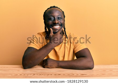 Handsome young black man wearing casual clothes sitting on the table looking confident at the camera smiling with crossed arms and hand raised on chin. thinking positive. 