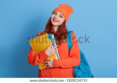 Friendly pleased young lady in cap carries rucksack, holds paper folder with papers, has tender smile, isolated over blue wall, dresses orange jumper, happy student being ready to study.