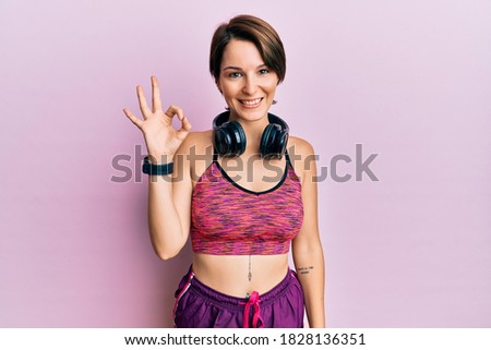 Young brunette woman with short hair wearing sportswear and headphones smiling positive doing ok sign with hand and fingers. successful expression. 