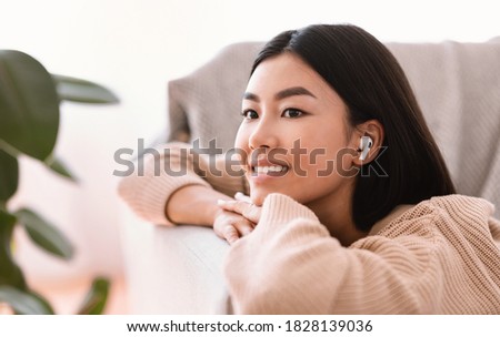 Enjoy The Music. Closeup portrait of calm asian woman wearing wireless earbuds listening to favorite song, relaxing on couch in living room. Smiling millennial lady lying on sofa and dreaming