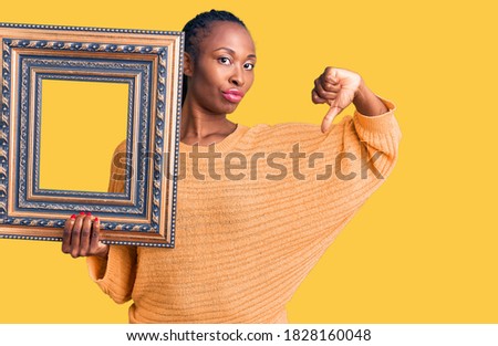 Young african american woman holding empty frame with angry face, negative sign showing dislike with thumbs down, rejection concept 