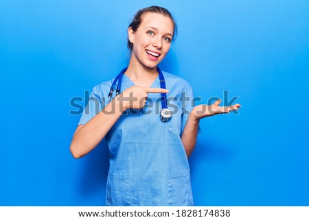 Young blonde woman wearing doctor uniform and stethoscope amazed and smiling to the camera while presenting with hand and pointing with finger. 
