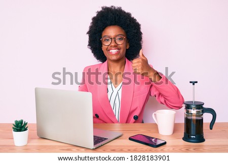 Young african american woman working at desk using computer laptop smiling happy and positive, thumb up doing excellent and approval sign 