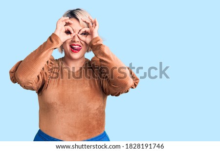 Young blonde plus size woman wearing casual sweater doing ok gesture like binoculars sticking tongue out, eyes looking through fingers. crazy expression. 