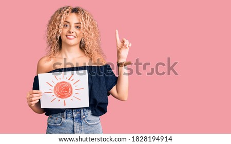 Young blonde woman with curly hair holding sun draw surprised with an idea or question pointing finger with happy face, number one 