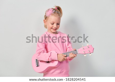 Cute caucasian little girl 4-5 year old in pink sweatshirt are playing the ukulele against gray studio background