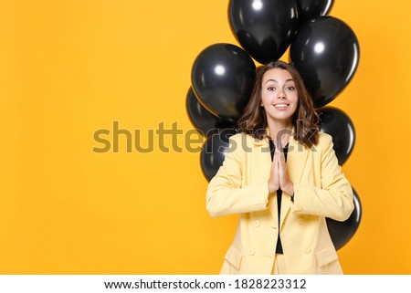 Pleading young woman 20s in suit jacket celebrating birthday holiday party with bunch of air ballons holding hands folded in prayer isolated on yellow background studio portrait. Black friday sale