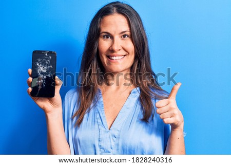 Young beautiful brunette woman holding broken smartphone showing cracked screen smiling happy and positive, thumb up doing excellent and approval sign