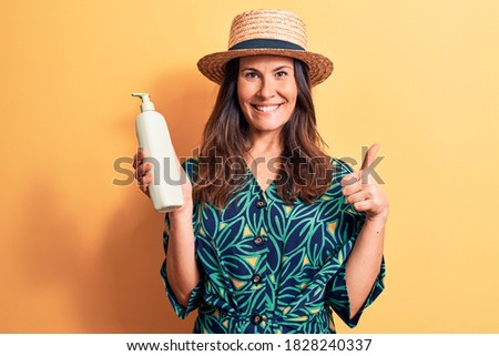Young beautiful brunette woman wearing summer hat holding bottle of sunscreen lotion smiling happy and positive, thumb up doing excellent and approval sign