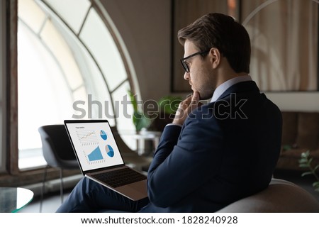 Side back view focused skilled young 30s male marketing specialist in suit analyzing graphs and charts in electronic research report on computer, developing sales growth strategy alone in office. Royalty-Free Stock Photo #1828240928