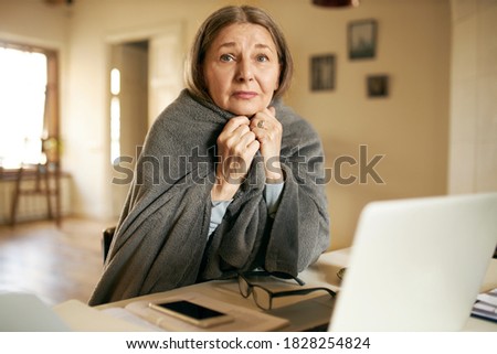 Portrait of unhappy elderly woman pensioner with mournful painful look feeling cold wrapping herself in warm plaid, receiving online medical help via video conference call with doctor using laptop Royalty-Free Stock Photo #1828254824