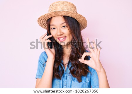 Young beautiful chinese girl wearing summer hat having conversation talking on the smartphone doing ok sign with fingers, smiling friendly gesturing excellent symbol 