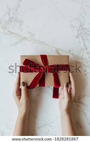 Female hands hold handmade paper gift box on marble background. Minimal festive Valentine's Day / Christmas / New Year celebration concept. Flat lay, top view.