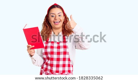 Young latin woman wearing apron holding recipe book smiling happy and positive, thumb up doing excellent and approval sign 