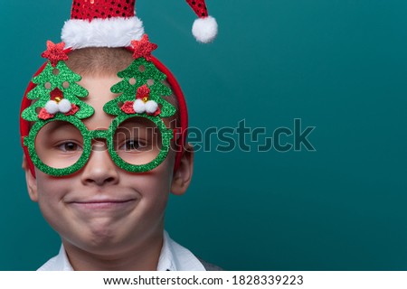 Portrait of happy cheerful boy wearing headband with Santa Claus Hat and funny glasses with Christmas trees posing on green wall. Merry Christmas and Happy New Year child.