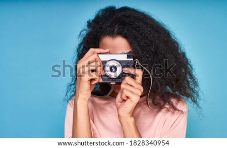 A close-up, curly brunette in a beige blouse, takes a picture on a retro camera, posing in the studio on a blue background.
