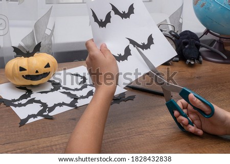child's hands are cutting from paper silhouettes of black bats, pumpkin with painted face lie near.  Getting ready for halloween party. Creative ideas concept.