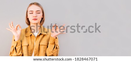 Mindful peaceful european woman meditates indoor, keeps hands in mudra gesture, has eyes closed, tries to relax after long hours of working, holds fingers in yoga sign, isolated on gray wall