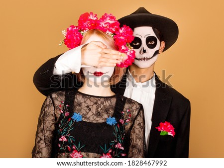Photo of calm relaxed couple ready for popular carnival, apply spooky makeup, man close woman's eyes against yellow background. Mexican holiday, people and celebration concept. Dead around us