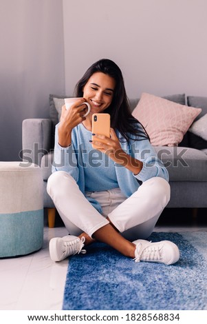 European fit brunette fashion blogger woman sits on floor in living room near sofa, wearing white jeans and blue sweather, and take photo selfie in mirror   