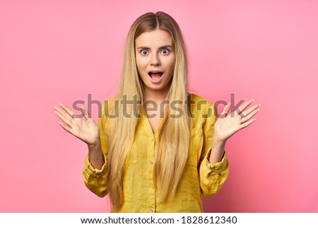 Surprised, frightened woman throws up her hands, looks at the camera, puzzled because she does not know the answer to a tricky question, isolated on a pink background. blonde