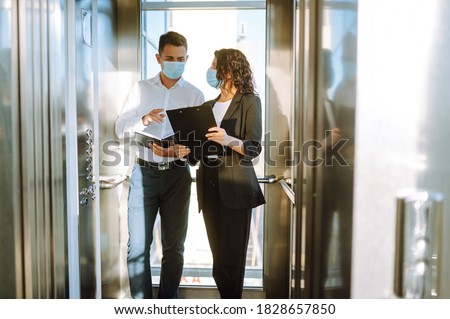 Business partners in protective sterile mask in the elevator. Colleagues talking while standing in elevator at  modern office. Teamwork during pandemic in quarantine city. Covid-19. Royalty-Free Stock Photo #1828657850