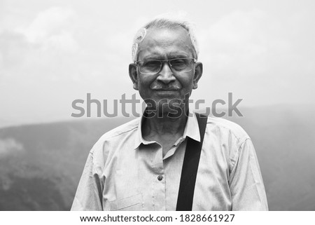 Monochrome portrait of an old man, wearing spectacles and blue shirt, standing alone in nature blurry mountain background. The elder person with a side bag on his shoulder, looking towards the camera.
