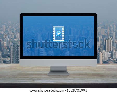 Play button with movie flat icon on desktop modern computer monitor screen on wooden table over office building tower and skyscraper in city, Business cinema online concept