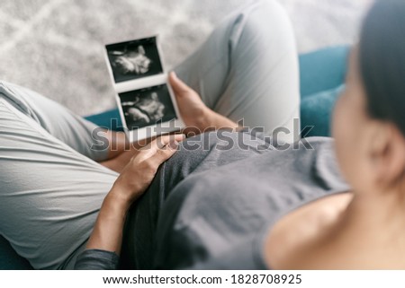 Pregnant woman relaxing on the sofa at home and watching ultrasound images of her baby, motherhood and pregnancy concept