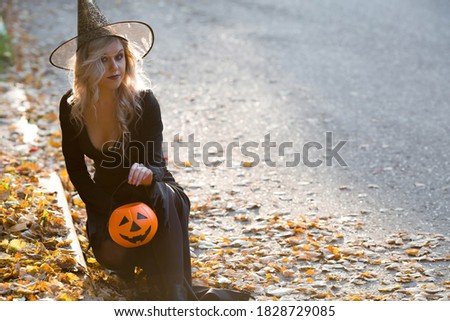 Halloween Witch with pumpkin. Beautiful young woman in a witch costume on the street. Halloween decor. Copy space.