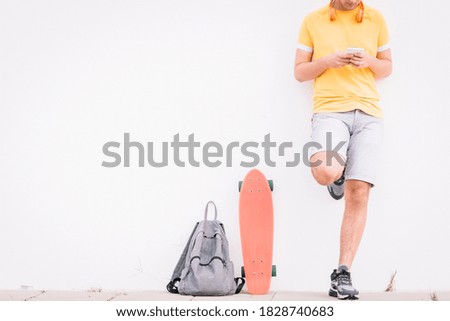 Teenager waiting leaning on the wall in a yellow shirt, with a skateboard and backpack.