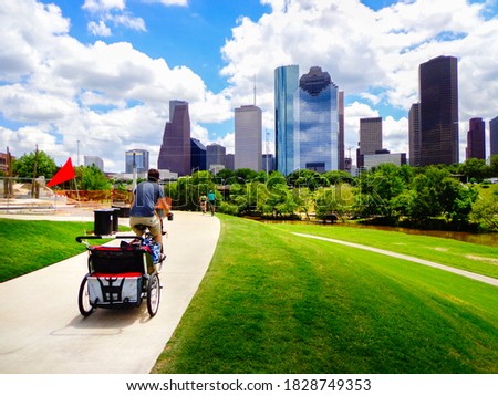 Young man riding bike with baby carriage attached on paved trail in Houston park (view of river and skyline of downtown Houston) - Houston, Texas, USA