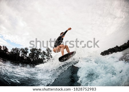 Active sportive woman in black wetsuit effectively jumps on surfboard on wave. Cloudy sky on the background