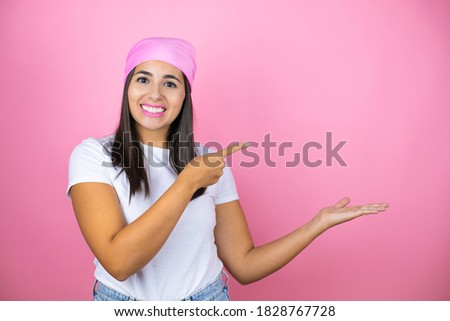 Young beautiful woman wearing pink headscarf over isolated pink background surprised, showing and pointing something that is on her hand