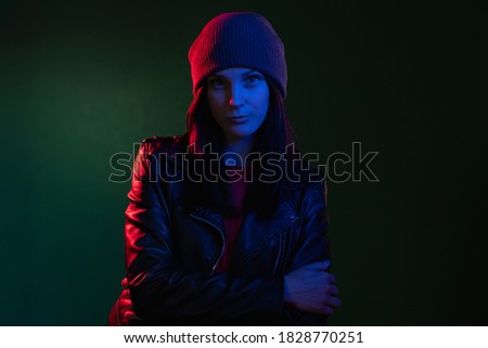 Night portrait. Neon light. Female power. Gender equality. Rebellious woman in hat leather biker jacket standing in red blue glow isolated on dark green copy space background.