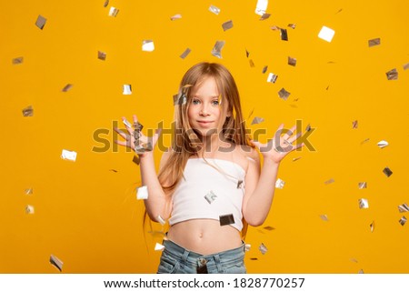 Festive celebration. Child party. Holiday fun. Birthday greeting. Excited happy blonde young girl enjoying confetti rain isolated on orange background.