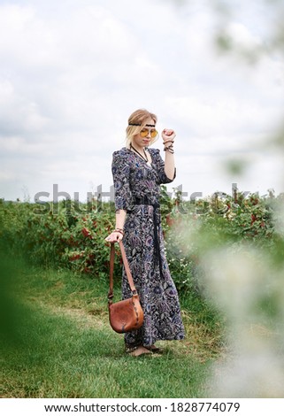 Young blond hippie woman, wearing grey boho style dress and yellow sunglasses, holding brown leather purse, standing on green field, posing for picture on sunny summer day. Inner freedom concept.